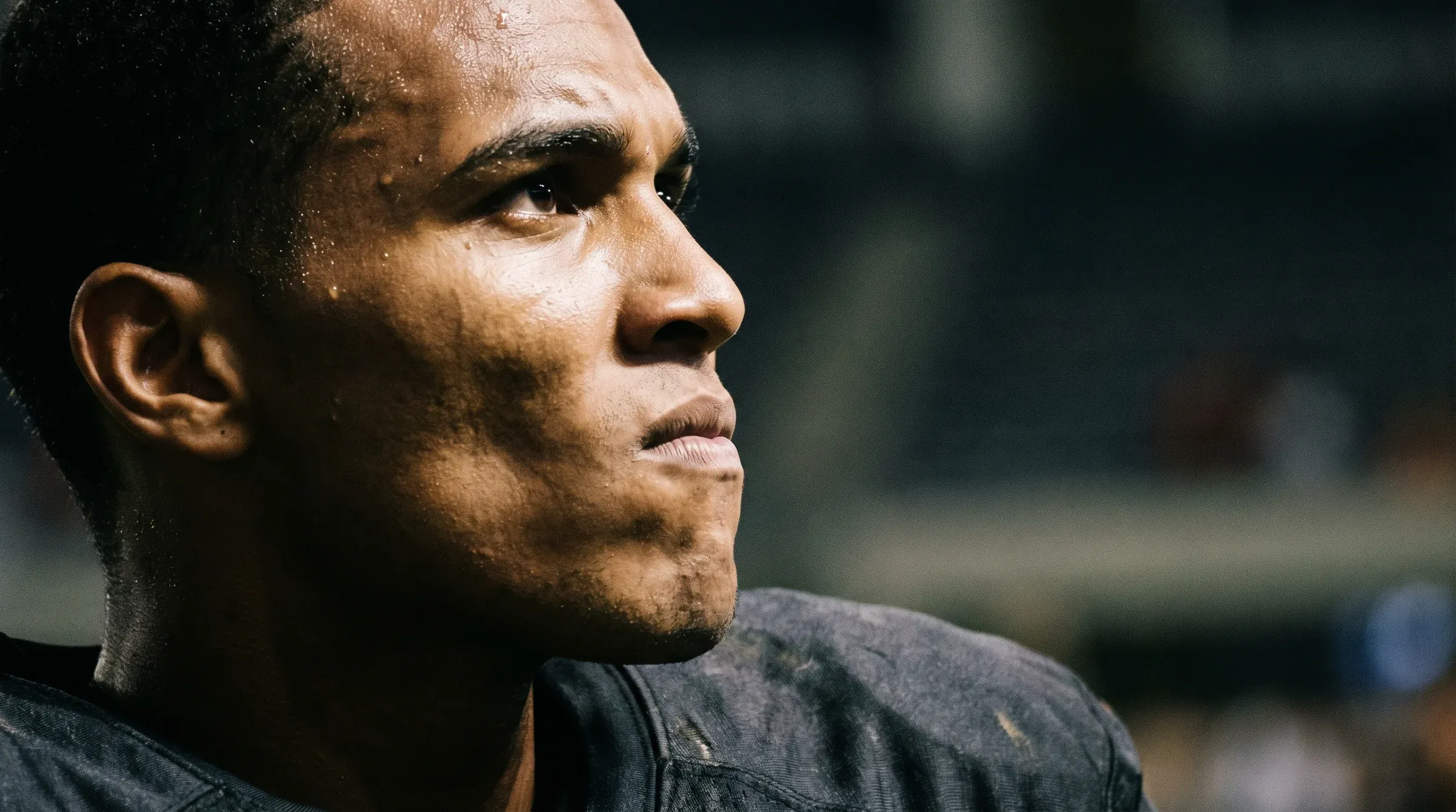 Tight photoreal portrait of a sweating Black athlete in dark gear looking up into the stadium lights, droplets on his skin glowing in the soft floodlight haze