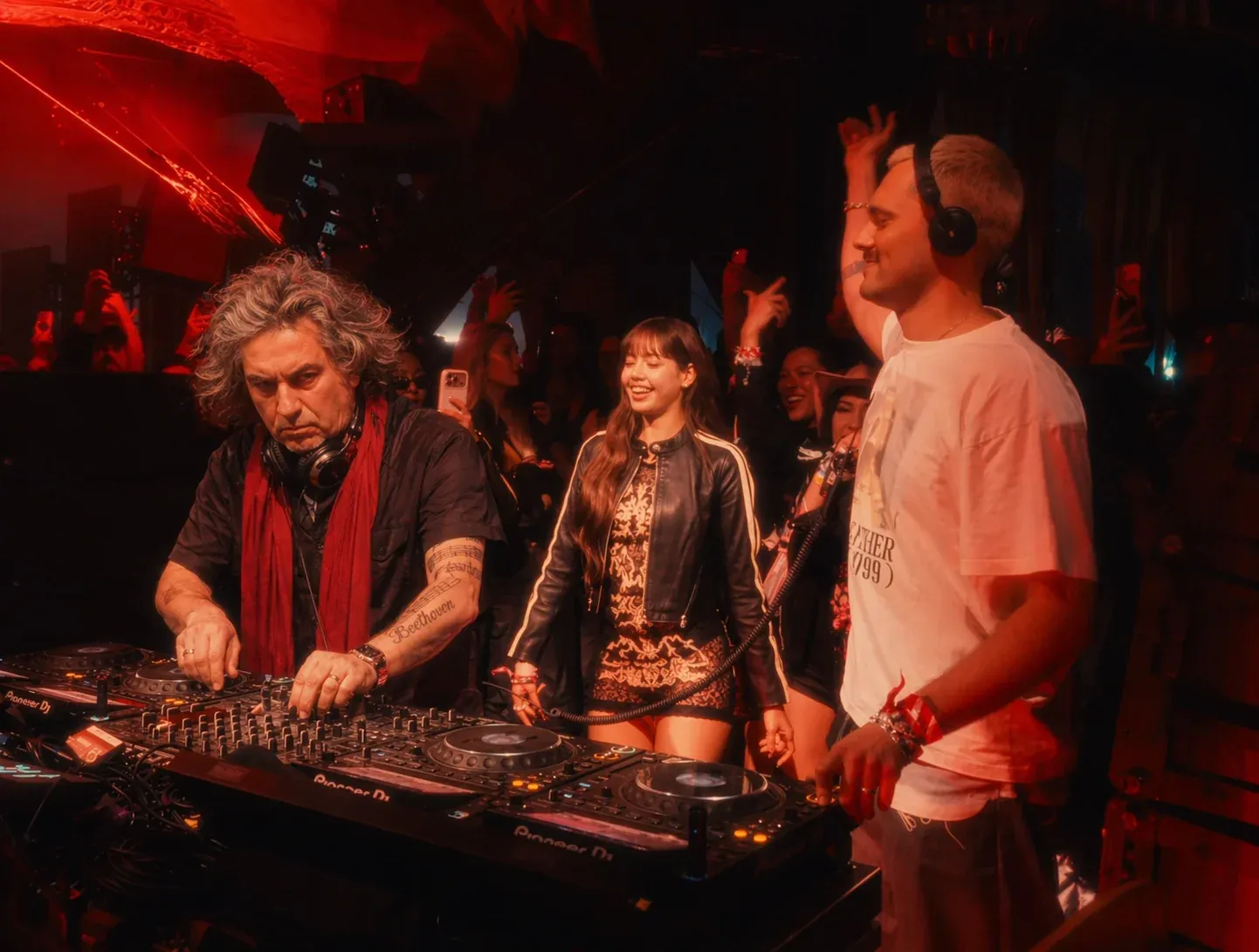 Long-haired Beethoven look-alike DJing at a packed festival stage between two younger DJs in white shirts under red and pink stage lights, dancing crowd in the foreground