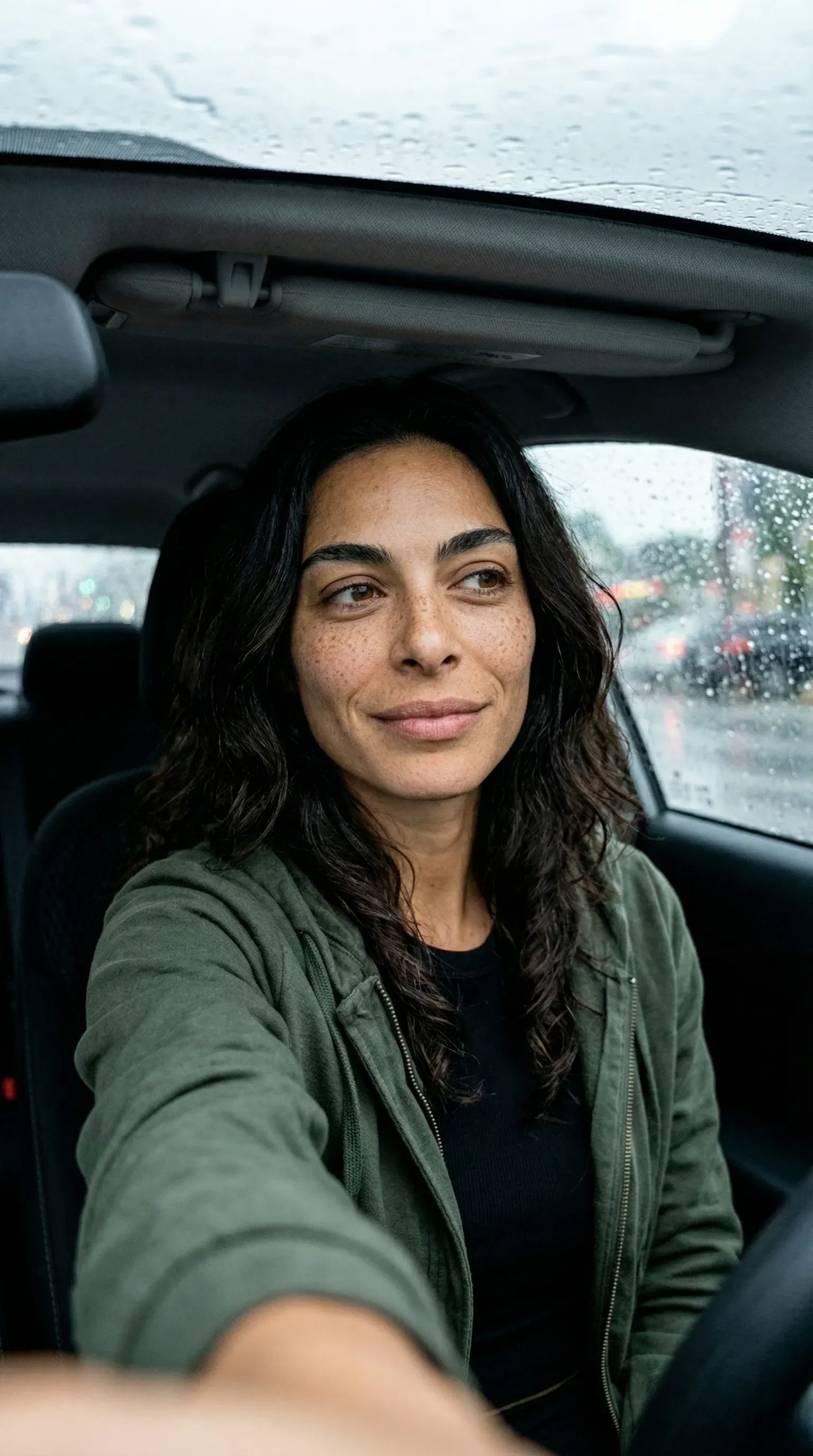 Selfie-style portrait of an olive-skinned woman with dark wavy hair in an olive green jacket sitting in a car on a rainy day with raindrops on the windshield, soft natural smile