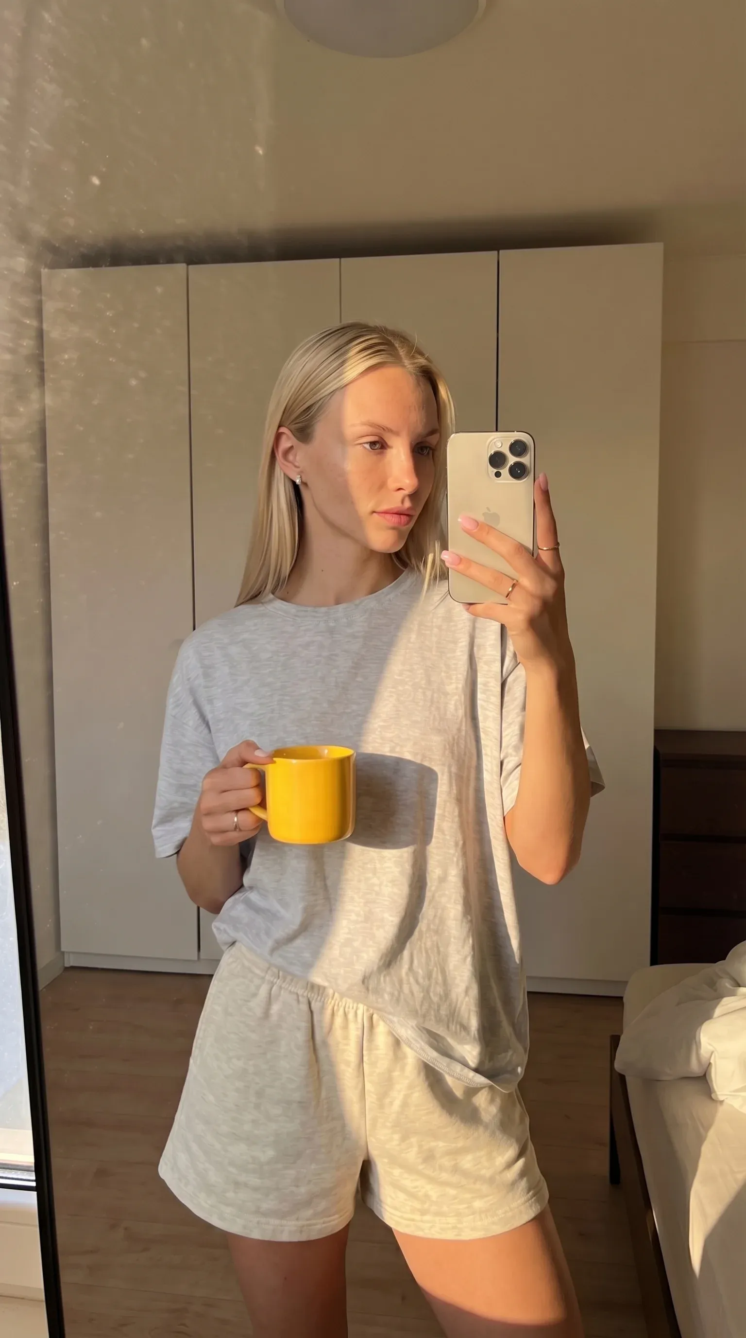 AI influencer Aleksia mirror selfie - young blonde woman in oversized grey t-shirt and beige shorts holding a yellow mug and iPhone, white wardrobe and corner of a bed behind her