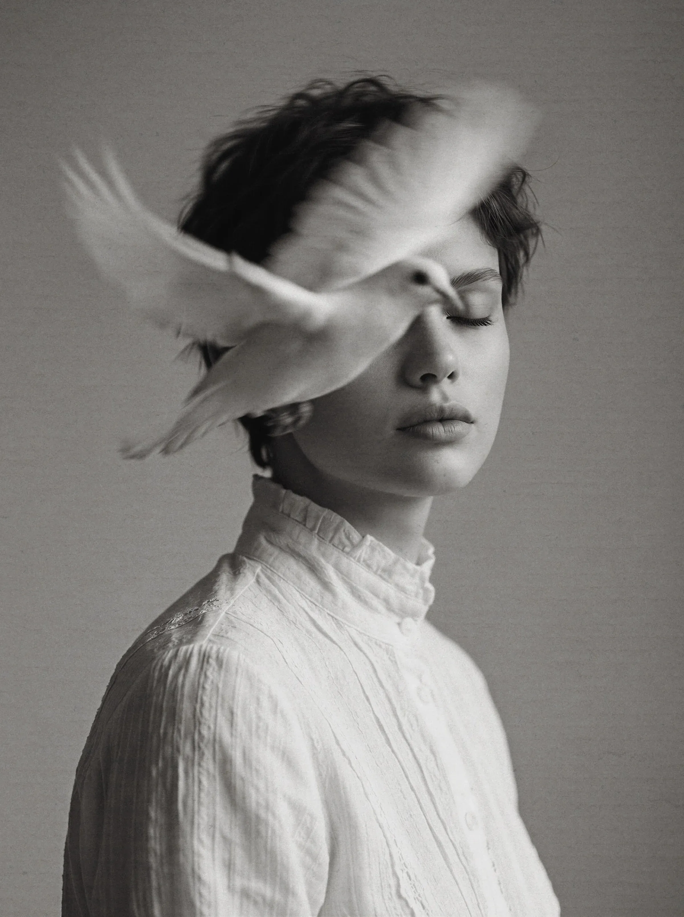 Black and white fine-art portrait of a short-haired woman in a high-collar white Victorian blouse with a white dove flying across her face and a dove wing covering her eyes