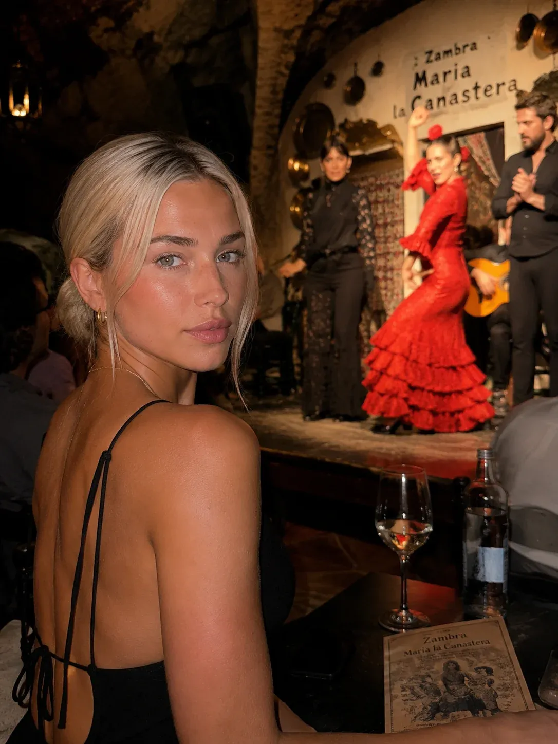 AI-generated portrait of a blonde woman in a black tied-back dress sitting at a candlelit Zambra Maria la Canastera tablao in Spain while a red-dressed flamenco dancer performs with a guitarist behind her