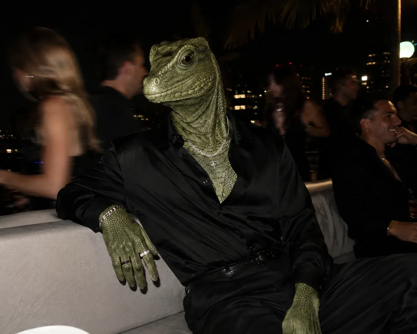 AI-generated photoreal lizard-headed humanoid in black silk pyjamas sitting on a rooftop lounge sofa at night with palm trees and blurred partygoers behind, taking the place of an AI influencer character