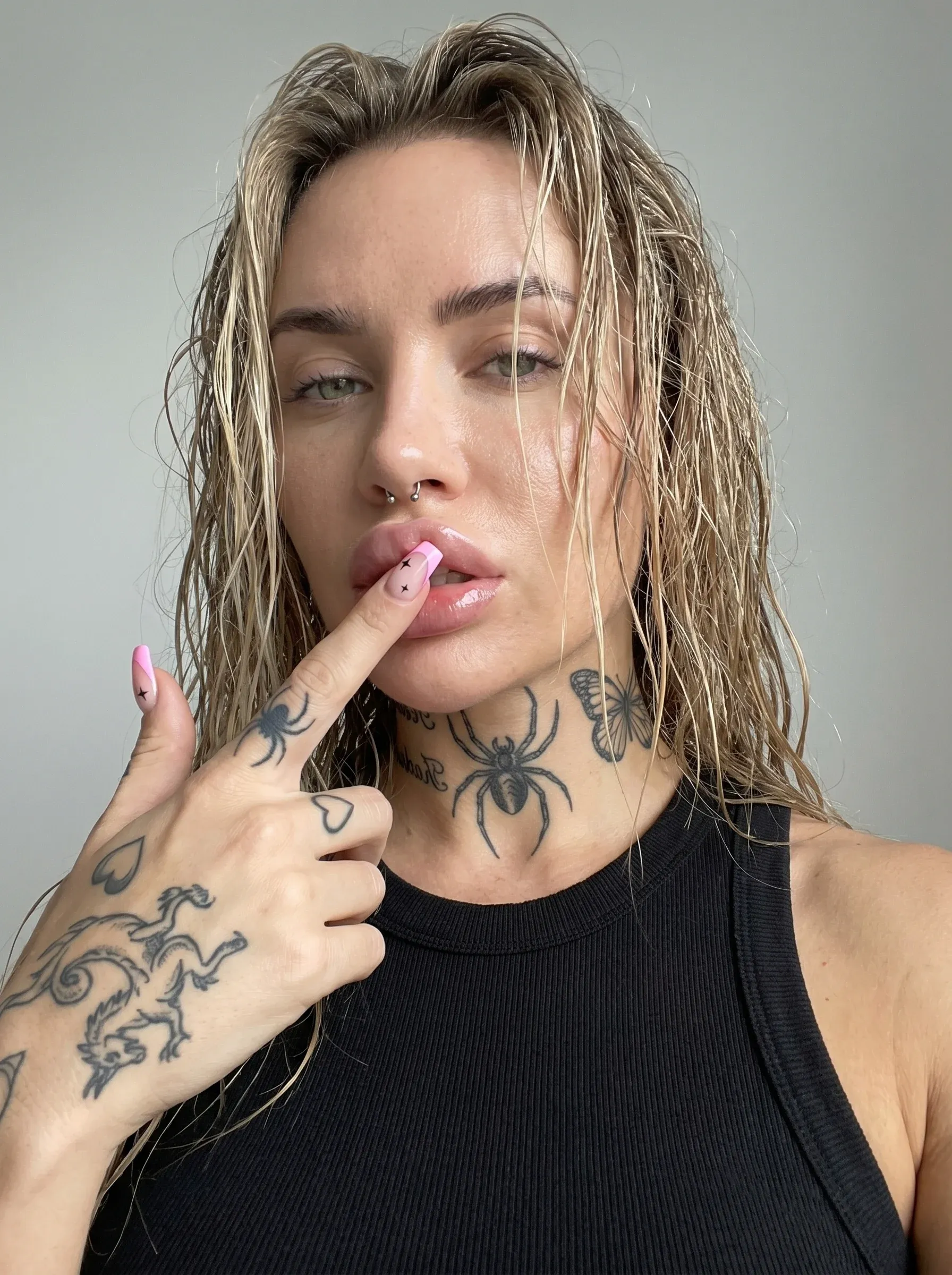 AI selfie-style portrait of a blonde woman with wet beach-hair touching her lower lip with a pink-painted finger, multiple spider and dragon tattoos visible across her neck, chest and hand