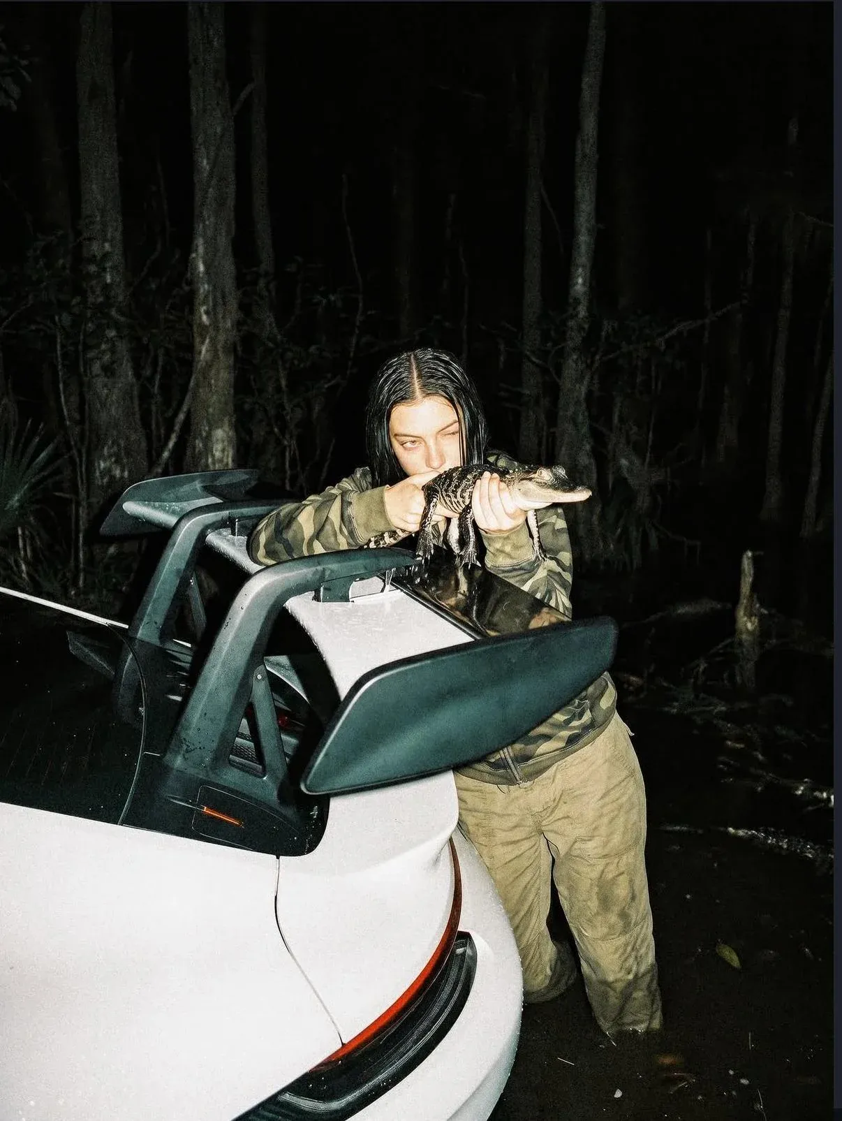 Moody night photo of a person in army-pattern jacket holding a small alligator across the back spoiler of a white sports car in a misty swamp forest
