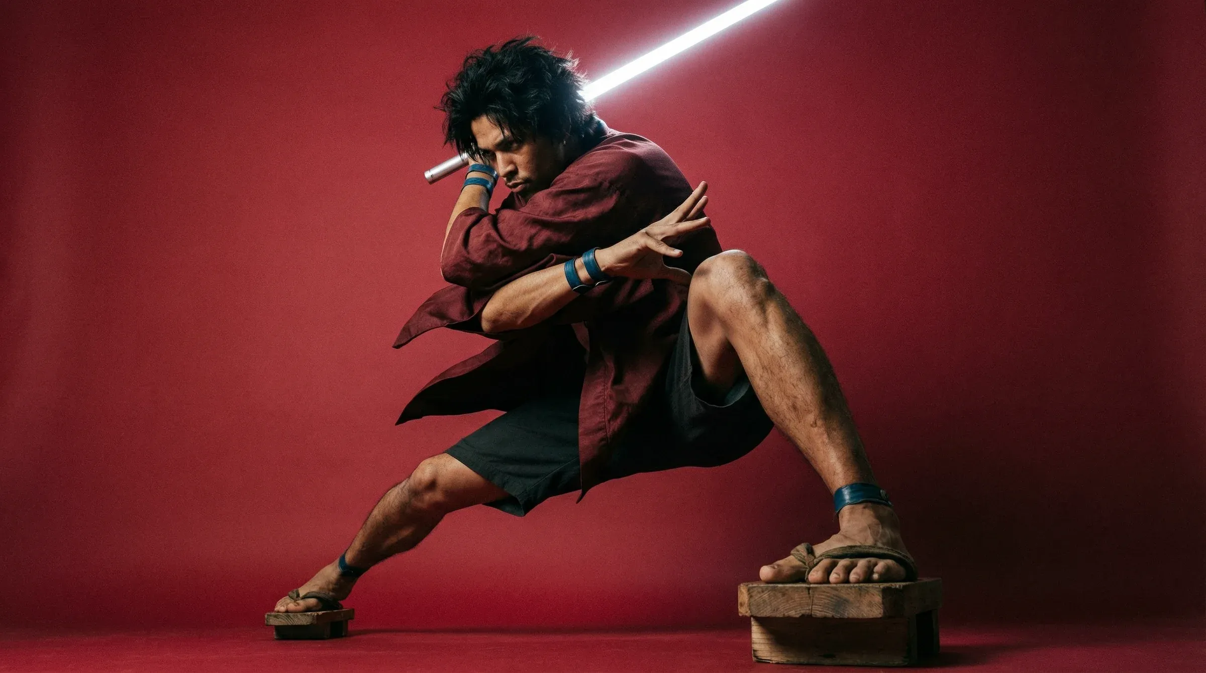 Cinematic studio portrait of a barefoot man in a dark red robe and wooden geta sandals in dramatic samurai stance brandishing a white lightsaber against a crimson backdrop