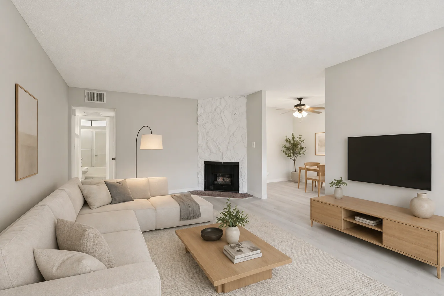 The same Park Providencia living room virtually staged by GPT Image 2.0 with a large L-shaped beige fabric sectional sofa facing the fireplace, a low wood coffee table on a soft cream area rug, a minimalist wood TV console on the right wall, an arc floor lamp near the sofa, a small olive tree in the dining alcove behind, and warm Scandinavian modern styling, with the original architecture, fireplace, walls, floors, and proportions preserved exactly