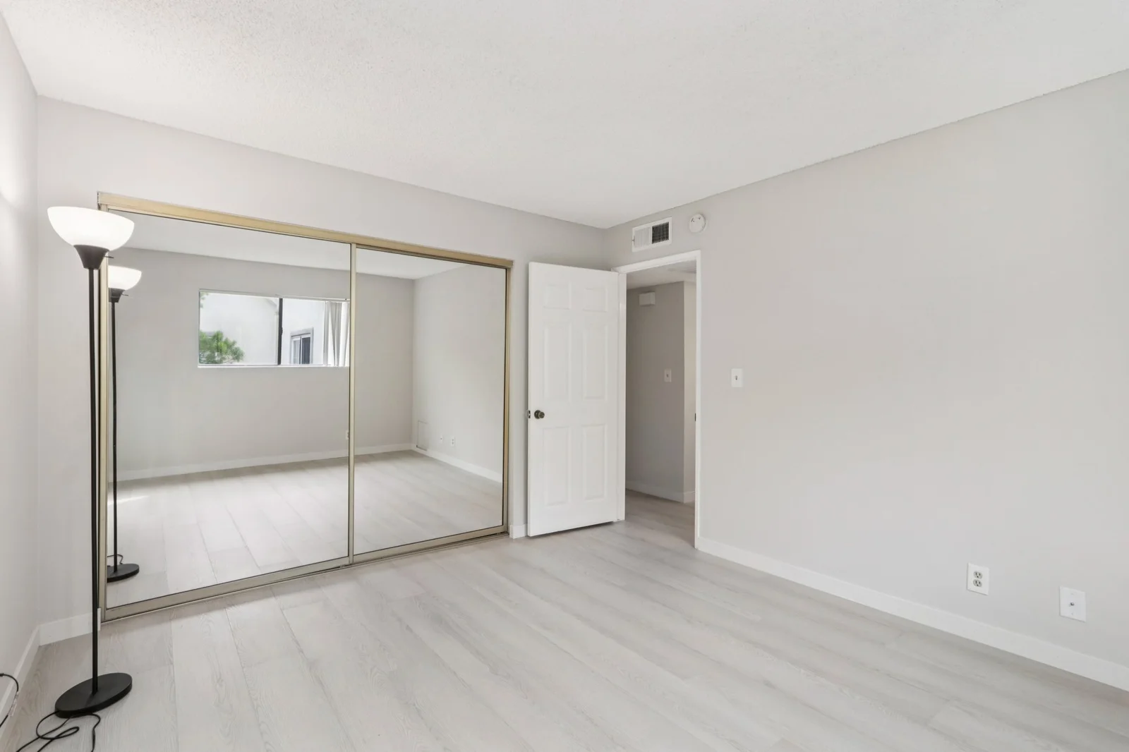 Empty bedroom photo from a publicly listed Park Providencia apartment in Burbank, California, sourced from Zillow, showing white walls, light wood floors, a tall floor lamp on the left, mirrored sliding closet doors that reflect the room, an open white interior door leading to a hallway, and an empty wall on the right