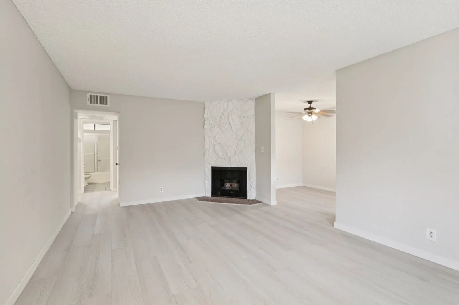Empty living room photo from a publicly listed Park Providencia apartment in Burbank, California, sourced from Zillow, showing white walls, light wood floors, white stone fireplace in the center, ceiling fan in the dining area, and a hallway leading to a bathroom on the left